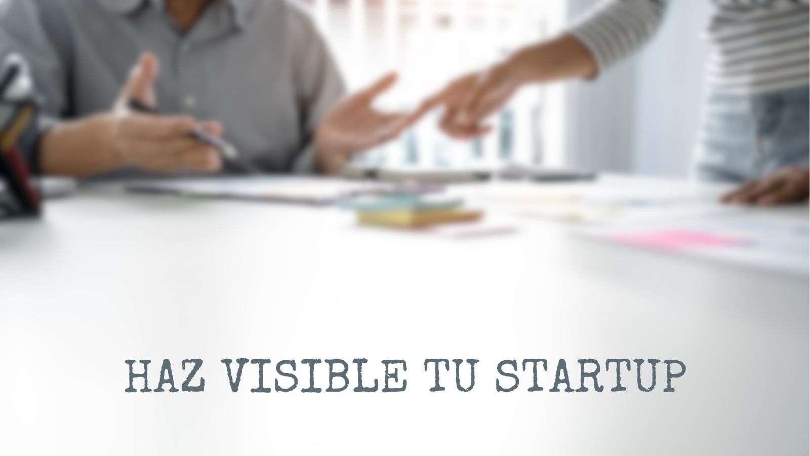 Dos personas en una reunión de negocios discutiendo estrategias sobre una mesa blanca con el texto "HAZ VISIBLE TU STARTUP".